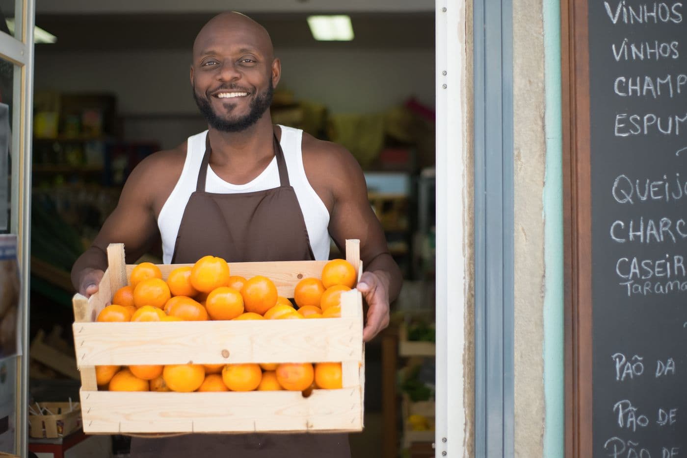 Comerciante sorrindo na porta da mercearia, com uma caixa de laranjas